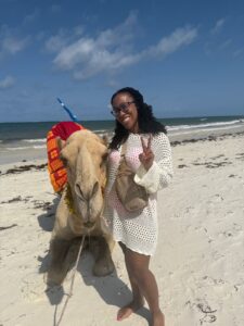 A woman stands next to a camel on the beach. 
